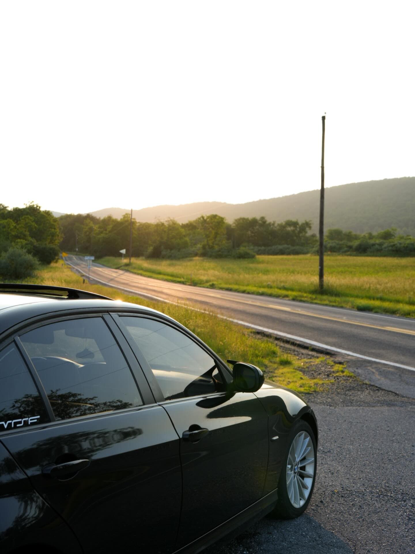 Conrad Captures photograph: black BMW E90 on a rural road at golden hour, mountains and fields in the distance.